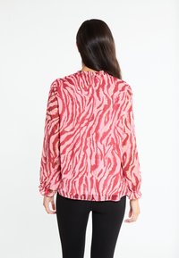 Blusa rosa trasparente con motivo a strisce zebra rosse, maniche lunghe con polsini elastici e scollatura arricciata. Indossata con leggings neri.