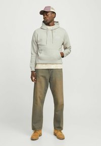Hellgrau Kapuzenpullover mit Logo, darüber ein gemustertes Hemd. Getragen mit lockeren, ausgewaschenen Jeans und hellbraunen Arbeitsschuhen. Abgerundet mit einer Mütze.