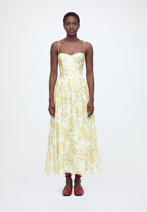 Γυναίκα φοράει κίτρινο φόρεμα με λουλουδένιο μοτίβο, χωρίς μανίκια και λεπτές τιράντες, και κόκκινασα γυναικεία παπούτσια με κορδόνια στον αστράγαλο, στέκεται μπροστά σε λευκό φόντο.