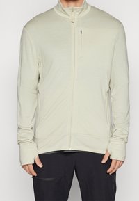 Chaqueta de cremallera beige claro con mangas largas, cuello alto y un bolsillo frontal. Textura suave, diseño aerodinámico y silueta ajustada.