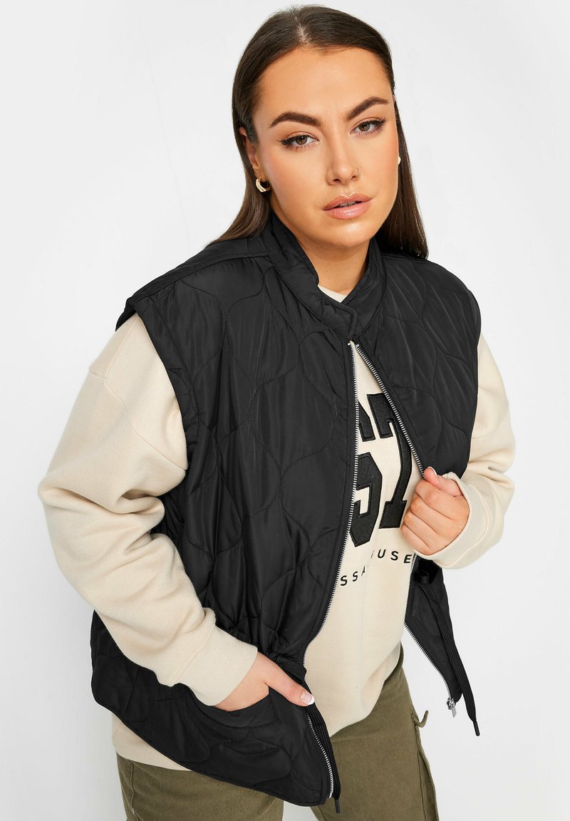 Svart vadderad väst med hög krage, framsida med dragkedja och sidofickor, bärs över en beige sweatshirt. Tyget ser mjukt och slätt ut.