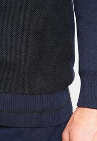 Marinblå tröja med en texturerad svart panel, ribbad kant och ärmar, med en slät yta. Sömmarna följer den böjda kanten.