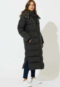 Kvinde iført en lang sort puffjakke med hætte, sidelukning, hænderne i lommerne, mørkeblå jeans og hvide sneakers, stående sidelæns.