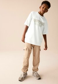 Hvid oversized t-shirt med trykt tekst, beige cargo bukser med sidelommer, og grå sportsko med hvide snørebånd.