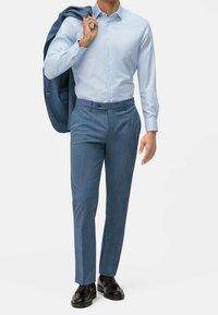 Chemise bleu clair à col français, associée à un pantalon bleu foncé ajusté et des chaussures noires, tenant une veste assortie posée sur une épaule.