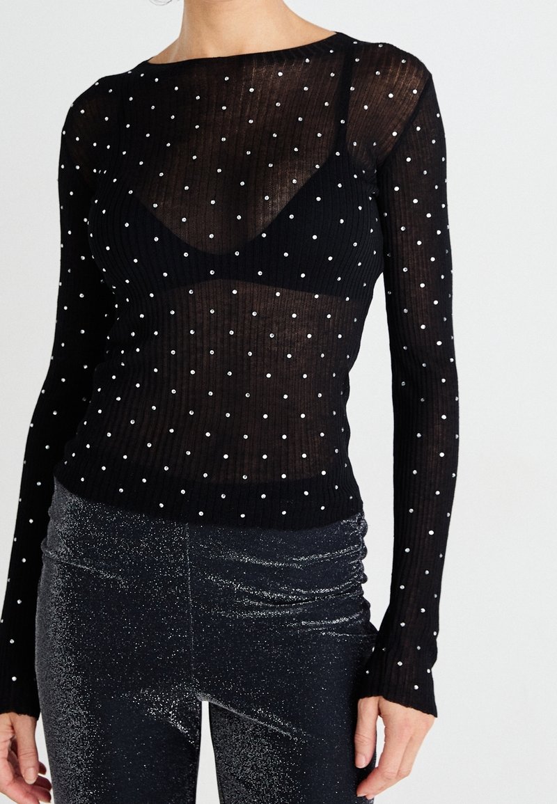 Haut transparent noir à manches longues avec des pois blancs porté sur un soutien-gorge noir, associé à un pantalon noir brillant taille haute.