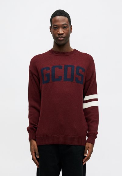 Burgunderrød strikkegenser med "GCOS" i marineblått, med hvite striper på ermene. Rund halslinning og ribbestrikkede mansjetter og nederkant.
