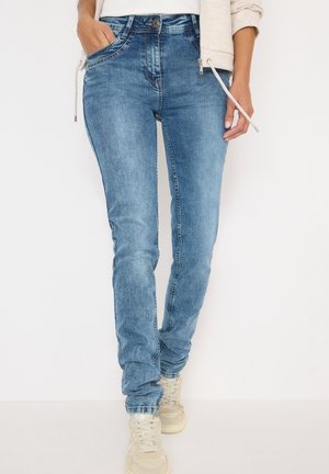 Jeans de mezclilla azul descoloridos con un corte ajustado, diseño de cinco bolsillos y cintura de altura media. Combinados con zapatillas en color beige claro.