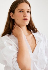 Bílá košile s vypouklými rukávy a nízkým výstřihem. Modelka má dlouhé hnědé vlasy, jednu ruku drží na krku a má na sobě tenký zlatý náramek.