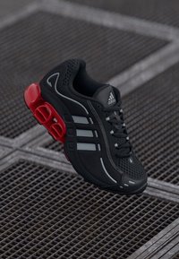 Crna sportska cipela s crvenim detaljima, gornjištem od mreže, tri bijele pruge i jastučićima za amortizaciju u potplatu, s uzorkom na teksturi.