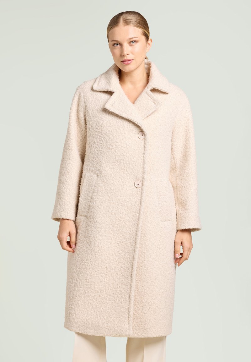 Abrigo de lana color crema con un acabado suave y texturizado, cuello con muesca, dos botones y bolsillos laterales. Mangas largas y diseño a la altura de la rodilla.