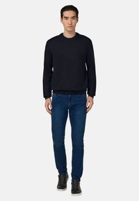 Zwart gemêleerd sweater, blauwe slim-fit jeans, zwarte sneakers met witte zolen. Model staat rechtop tegen een effen achtergrond.