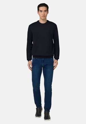 Schwarzer strukturierter Pullover, blaue Slim-Fit-Jeans, schwarze Sneakers mit weißen Sohlen. Das Modell steht aufrecht vor einem schlichten Hintergrund.