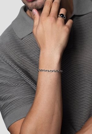 Silver chain bracelet on wrist, featuring interlocking links. Gray polo shirt with textured pattern, short sleeves, and ribbed trim.