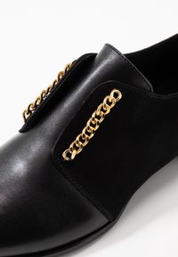 Chaussure noire en cuir lisse et en velours, agrémentée de détails en chaîne dorée sur les côtés. Design élégant et moderne.