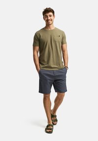 Camiseta de manga corta verde oliva con bolsillo, pantalones cortos azul marino y sandalias verdes. Ajuste casual y relajado con un diseño simple.