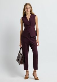 Gilet bordeaux su misura con un unico bottone dorato, abbinato a pantaloni slim-fit coordinati e borsa marrone. Tacchi in suede completano il look.