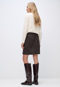 Mini jupe en daim brun, chemise à manches longues crème, et bottes brunes vernies à genoux avec boucles, vues de dos.