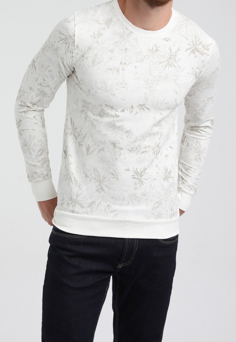 Homme portant un pull blanc à manches longues avec un motif géométrique et floral subtil, associé à un jean noir, debout devant un fond uni.
