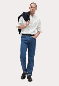 Camisa de rayas claras con mangas enrolladas, jeans de mezclilla azul clásicos, cinturón negro y zapatos de vestir negros. Suéter caído sobre el hombro.