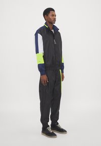 Black windbreaker with mesh accents in navy and neon green, paired with matching black joggers. Features elastic cuffs and a front zipper.
