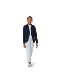 Blazer azul-marinho com dois botões, calças cinza claras e uma t-shirt gráfica branca. Modelo usa ténis brancos, caminhando sobre um fundo branco.