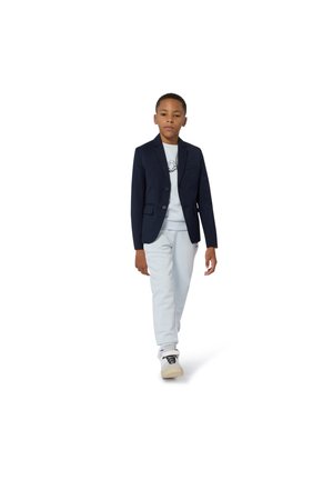 Blazer navy à deux boutons, pantalon gris clair et t-shirt graphique blanc. Le modèle porte des baskets blanches, marchant sur un fond blanc.