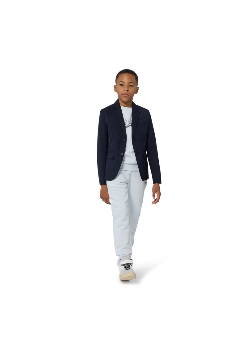 Blazer azul-marinho com dois botões, calças cinza claras e uma t-shirt gráfica branca. Modelo usa ténis brancos, caminhando sobre um fundo branco.