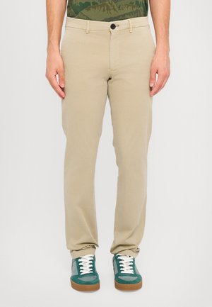 Mann trägt beige Hose und grün-graue Sneaker und steht vor weißem Hintergrund.
