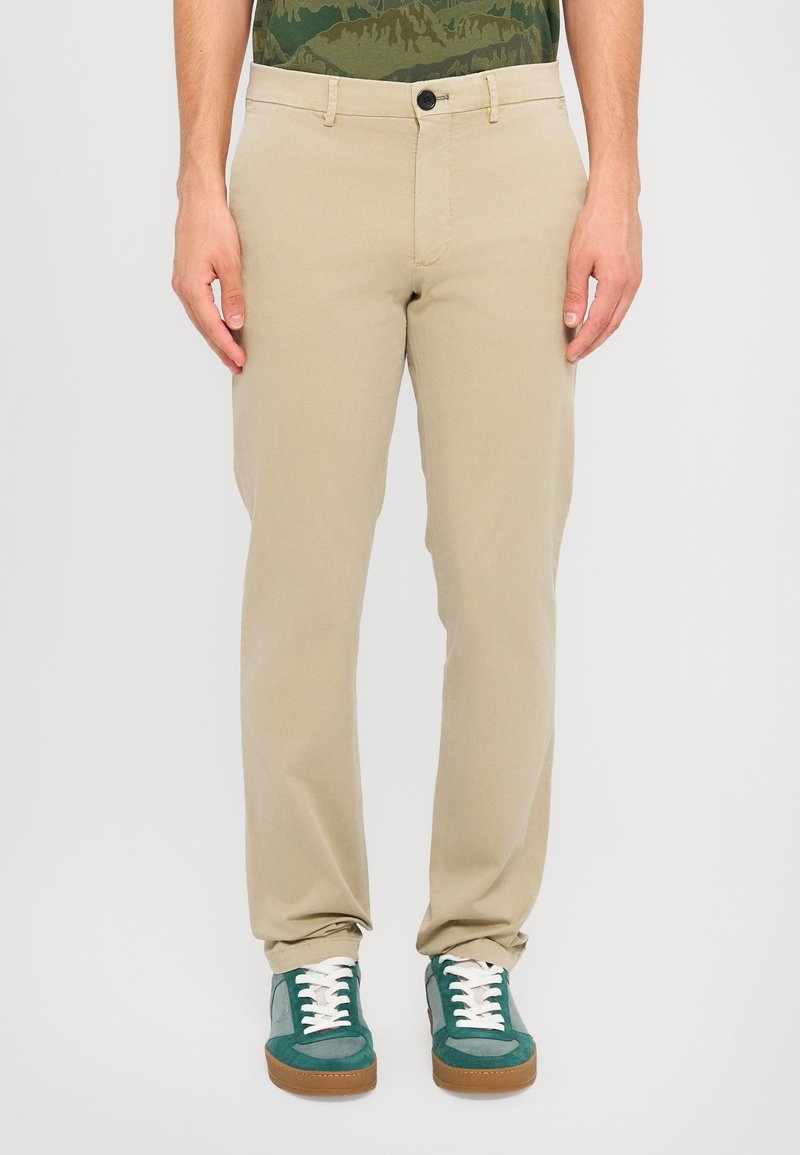 Homme portant un pantalon beige et des baskets vertes et grises, debout sur fond blanc.
