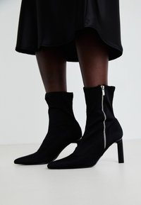Botas negras hasta el tobillo con un cuerpo de tela elástica, punta afilada y cierre lateral de cremallera, sobre un tacón fino y alto.