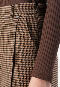 Detailný záber na osobu oblečenú v nohaviciach so vzorom houndstooth v hnedej a čiernej farbe, s predným zipsom a gombíkom, v kombinácii s hnedým dlhým rukávom s jemným ryhovaním.