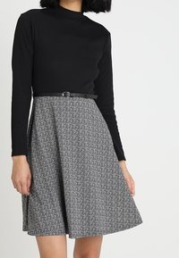 Svart långärmad topp kombinerad med en knälång kjol med hundtandsmönster i svart och vitt, accentuerad med ett smalt svart skärp.