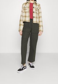 Skuren rutig jacka i beige och gult, röd topp, olivgröna cargobyxor med sidofickor och svarta sneakers med vit sula.