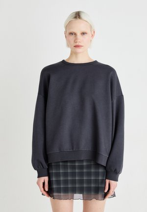 Sweatshirt gris avec une coupe décontractée, des poignets et un ourlet côtelés, superposé sur une jupe à carreaux noir et vert avec un ourlet en volants.