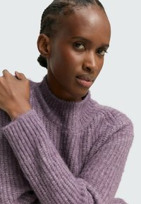 Fioletowy sweter przędzy z wysokim kołnierzem, o prążkowanej strukturze i luźnym kroju. Miękki materiał z widocznymi detalami szycia.