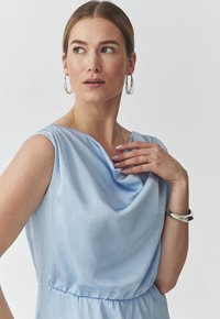 Světle modré šaty s volně draped výstřihem a elastickým pasem. Hladká textura, bezrukávový design a jemný lesk jsou hlavními rysy.