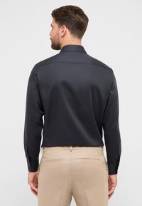 Camicia nera a maniche lunghe con una texture liscia, colletto strutturato e polsini con bottoni, abbinata a pantaloni beige chiaro con tasche posteriori.