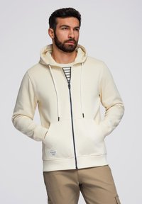 Felpa crema con zip, coulisse bianche, tasche frontali e un'etichetta sul lato. Il design a righe sotto la zip aggiunge un tocco di dettaglio.