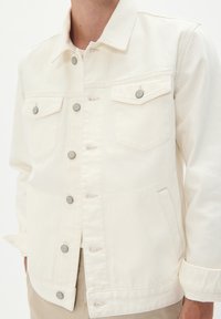 Jachetă din denim alb cu guler clasic, închidere cu nasturi, două buzunare pe piept și nasturi de ton argintiu. Material texturat cu o finisare curată.