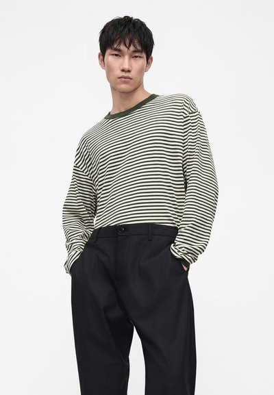 Jeune homme aux cheveux courts et noirs portant une chemise à manches longues rayée vert et blanc et un pantalon noir, les mains dans les poches.