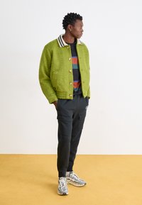 Zöld textúrájú bomberdzseki csíkos nyakkal, egy fekete és többszínű csíkos póló felett viselve; sötét cargo nadrággal és sportcipővel párosítva.