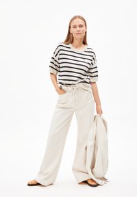 Gestreepte oversized t-shirt in crème en marineblauw, gecombineerd met beige wijde broek. Model houdt een bijpassende jas vast en draagt bruine sandaal.