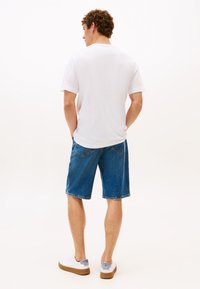 Fehér rövid ujjú inget, kék farmer rövidnadrágot és fehér tornacipőt viselő férfi, zsebében tartott kézzel, háttal állva.