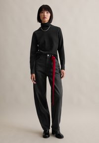 Fekete garbós pulóverben, laza fekete farmernadrágban, hosszú piros övvel megkötve, fekete bokacsizmában álló nő egyszínű háttér előtt.