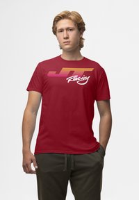 Röd bomull t-shirt med en flerfärgad grafik av "JT Racing" med horisontella linjer i rosa och orange, korta ärmar och rund hals.