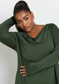 Mörkgrön långärmad topp med en lös hals, tillverkad av stretchigt material. Har långa ärmar med tumhål och en avslappnad passform.