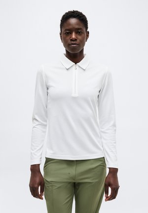 Naine seisab ühevärvilise tausta ees, seljas valge pikaveoline topp lukuga kraega ja oliivrohelised püksid, suunaga edasi neutraalse ilmega.