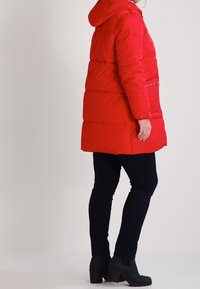 Červená péřová bunda s kapucí, se zipovým zapínáním a vertikálními švy. Kombinována s tmavými úzkými džíny a černými kotníkovými botami s blokovým podpatkem.