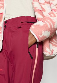Bordeauxfarvede tekniske bukser med bælte, der har en lynlåslomme og en glat tekstur. Kombineret med en mønstret pink fleecejakke.